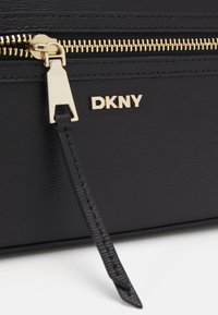Carteira de couro preto com fecho dourado, logótipo DKNY dourado, acabamento texturizado e puxador de fecho decorativo. Apresenta um design liso e estruturado.