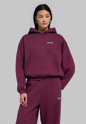 Femme portant un sweat à capuche violet WRSTBHVR et un pantalon de jogging assorti, debout devant un fond gris uni, regardant sur le côté.