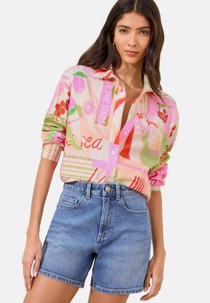 Woman wearing a multicolored, patterned long-sleeve shirt tucked into blue denim shorts, standing against a plain background.