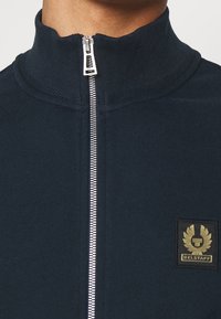 Marinblå zip-up jacka gjord av mjukt tyg; har en hög krage och en silverdragkedja. Märkbart guldlogom ärke på vänster bröst.