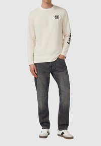 Beige overhemd met lange mouwen en een zwart logo op de borst, met de tekst "Discovery" langs de mouw, gecombineerd met donkergrijze jeans en witte sneakers.
