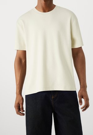 Mężczyzna w jednolitym, ecru T-shircie z krótkim rękawem i dekoltem okrągłym, w połączeniu z ciemnoniebieskimi dżinsami, stojący na białym tle.