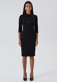 Donna che indossa un vestito nero aderente lungo fino al ginocchio, con maniche a tre quarti, chiusura frontale con bottoni, colletto, orecchini argentati e tacchi alti neri.