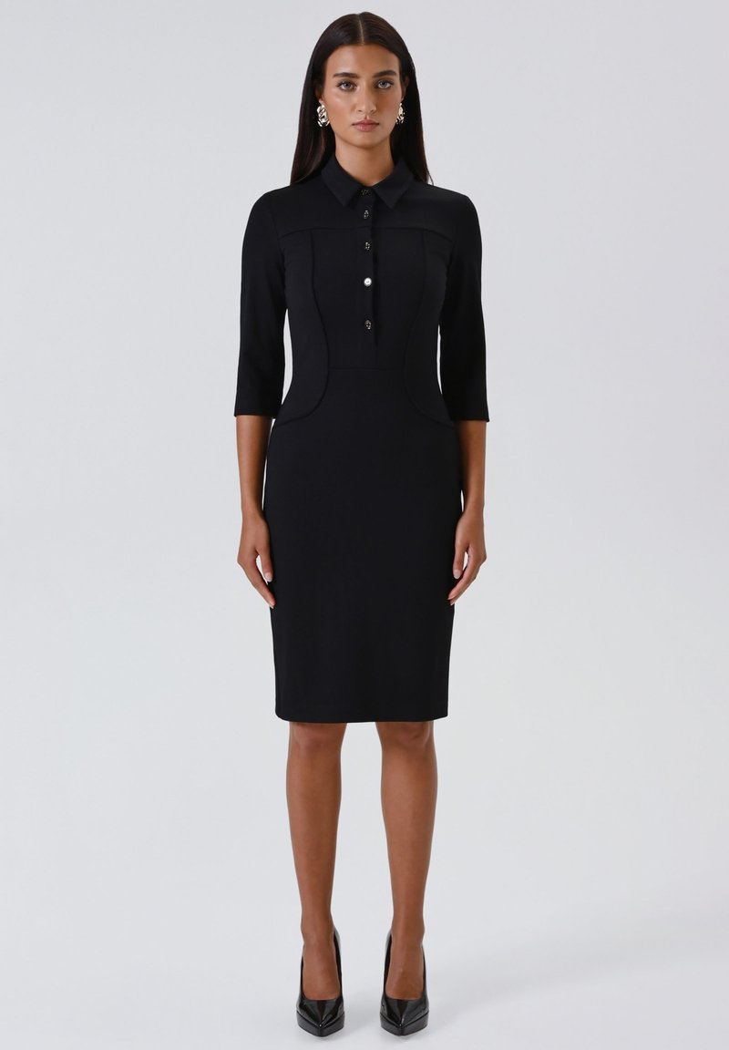 Donna che indossa un vestito nero aderente lungo fino al ginocchio, con maniche a tre quarti, chiusura frontale con bottoni, colletto, orecchini argentati e tacchi alti neri.