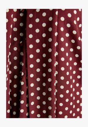 Bordo audums ar baltām punktiņām, kuram ir gluda tekstūra un salocīta dizaina, rādīdams konsekventu motīvu ar dažāda izmēra punktiem.