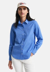 Camicia a maniche lunghe blu in tessuto con bottoni sul davanti e colletto. Cappellino da baseball bianco con testo rosso e blu, indossato con pantaloni bianchi.