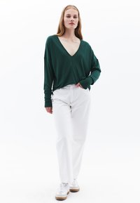 Grüner V-Ausschnitt-Pullover aus weichem Material, kombiniert mit weißen weiten Hosen und weißen Sneakers mit einem braunen Sohlen. Das Model steht vor einem weißen Hintergrund.