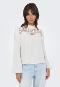 White long-sleeve top with lace detailing on the shoulders and chest, paired with light blue jeans. Soft fabric and relaxed fit.