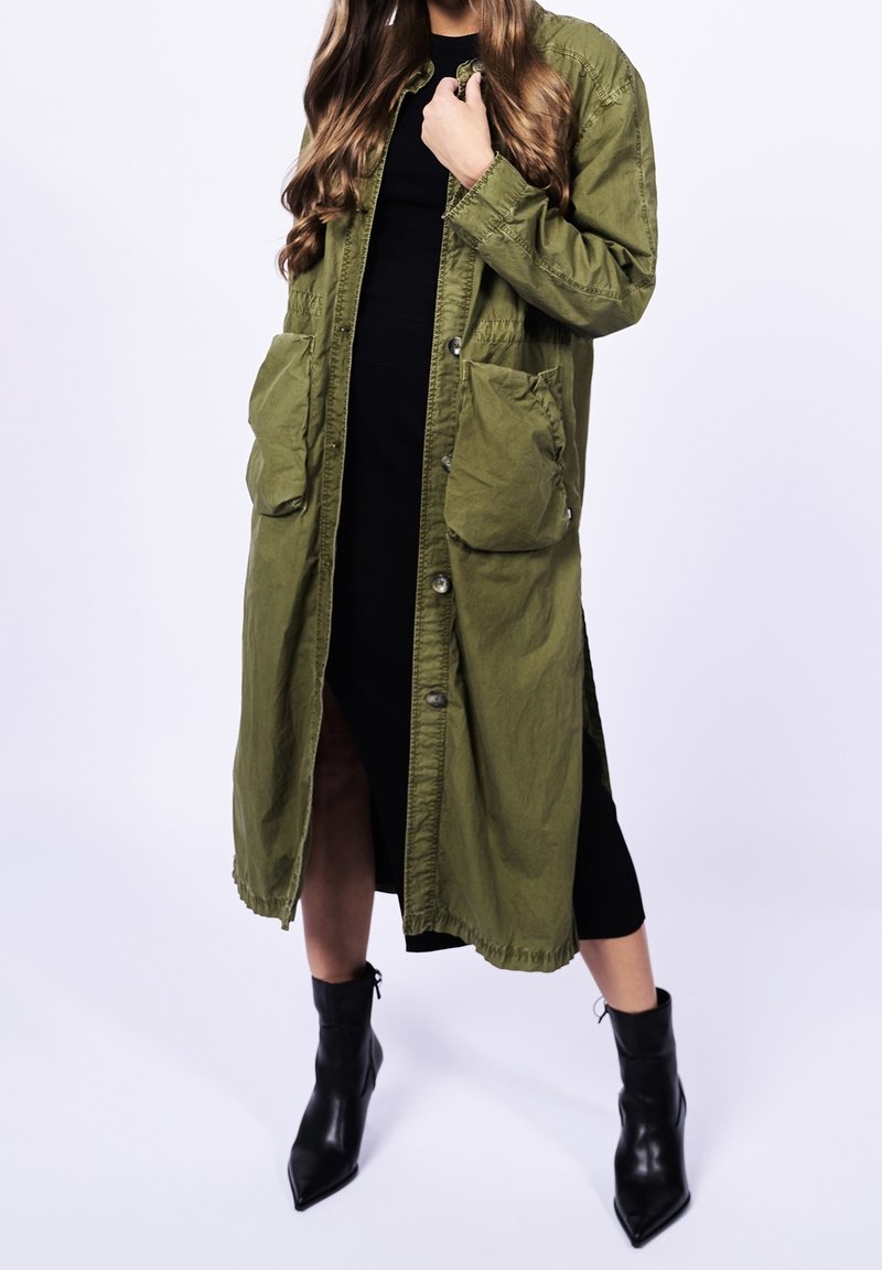 Femme portant un long manteau boutonné vert olive sur une robe noire, associé à des bottines noires à bout pointu, debout sur un fond blanc.