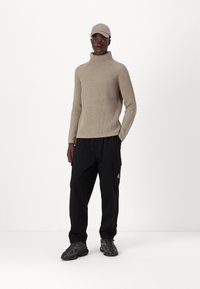 Beige, gerippter Rollkragenpullover mit langen Ärmeln, kombiniert mit schwarzen Cargo-Hosen und dunklen Sneakers. Inklusive einer beige Kappe.