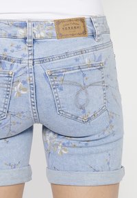 Shorts de mezclilla con un estampado floral en azul claro, con dobladillos enrollados y una etiqueta marrón en la parte trasera de la cinturilla. Cuenta con dos bolsillos traseros.
