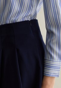 Camisa a rayas azul claro y blanca combinada con pantalones oscuros de pinzas de color navy, que presentan una textura suave y un discreto detalle de cremallera.