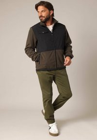 Fleece jack in zwart en olijfgroen met ritssluitingen, hoge kraag en elastische manchetten, gedragen met olijfgroene broek en witte sneakers.