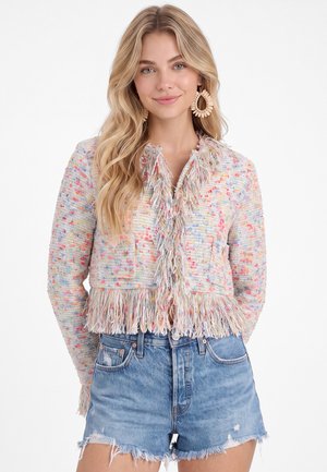Lyshåret kvinde med bølget hår har en flerfarvet frynsejakke og blå denimshorts på, smilende mod en hvid baggrund.