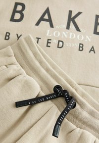 Sudadera de algodón beige con texto del logo en negro. Presenta una cintura elástica y un cordón negro con un patrón de nombre de marca repetido.