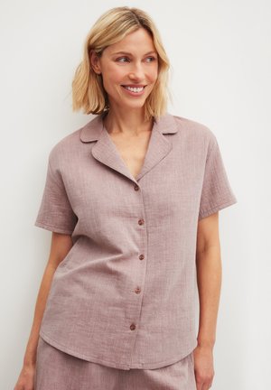 Smiling woman with blonde hair wearing a short-sleeve, buttoned, light mauve linen shirt and matching bottoms against a white background.