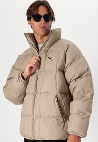 Giacca a vento beige con collo alto, chiusura con zip e tasche laterali. Presenta pannelli cuciti e un logo nero sul petto.