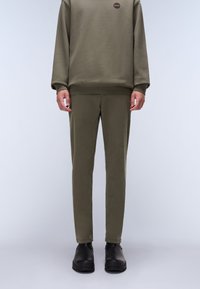 Man i en olivgrön sweatshirt och matchande slim-fit byxor med svarta ankelstövlar, stående mot en ljus platt bakgrund.