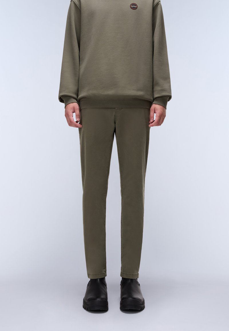 Hombre vestido con sudadera verde oliva y pantalones slim-fit a juego, con botas negras hasta el tobillo, de pie contra un fondo claro y liso.