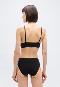 Γυναίκα με κοντά καστανά μαλλιά, που στέκεται γυρισμένη πίσω, φοράει μαύρο σουτιέν και εσώρουχα μπροστά σε απλό φωτεινό φόντο.