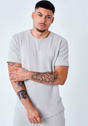 T-shirt gris à manches courtes avec une finition texturée, présentant un logo subtil sur la poitrine. Le modèle montre des bras tatoués et un arrière-plan neutre.