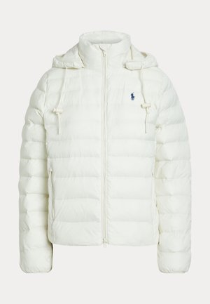 Γυναίκα στέκεται με τα χέρια στις τσέπες φορώντας κρεμ μπουφάν, μπεζ πουλόβερ με ψηλό λαιμό, φαρδιά παντελόνια, κάλτσες και λευκά αθλητικά παπούτσια.