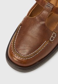Chaussure en cuir marron avec un design à bride en T, des accents cousus et un bout arrondi. La semelle est noire et légèrement surélevée.