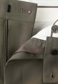 Gros plan d’un pantalon gris montrant un bouton, une fermeture éclair, des boutonnières et une doublure intérieure blanche avec des détails de couture.