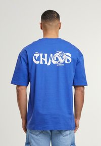 Blå bomulds t-shirt med korte ærmer, med stort hvidt "CHAOS" tekst på ryggen og en bølgeskabelon, afslappet pasform og rund hals.
