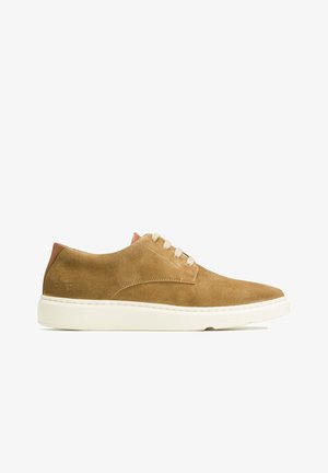 Chaussures à lacets en daim marron clair avec une semelle en caoutchouc blanche. Elles comportent un détail en cuir marron clair au talon et des lacets beige. Texture lisse dans l'ensemble.