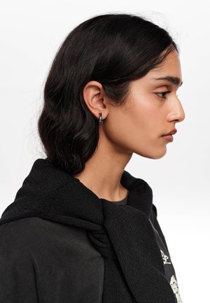 Joven mujer con camisa negra y suéter negro sobre los hombros, lleva un pequeño pendiente de aro colorido y mira hacia la derecha sobre un fondo blanco.