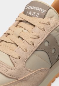 Gros plan sur une sneaker beige Saucony Jazz montrant les lacets, les panneaux en daim et tissu, ainsi que le logo sur la languette et le côté.