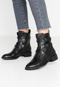 Botas de tobillo de cuero negro con múltiples correas con tachuelas, un zipper lateral y un tacón bajo. Combinadas con jeans grises desgastados.
