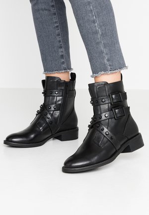 Bottines en cuir noir avec plusieurs sangles ornées de clous, une fermeture éclair latérale et un petit talon carré. Portées avec un jean gris effiloché.