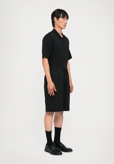 Polo noir à manches courtes, associé à un short noir à cordon de serrage. Porté avec des chaussures habillées noires et des chaussettes noires de longueur mi-mollet.