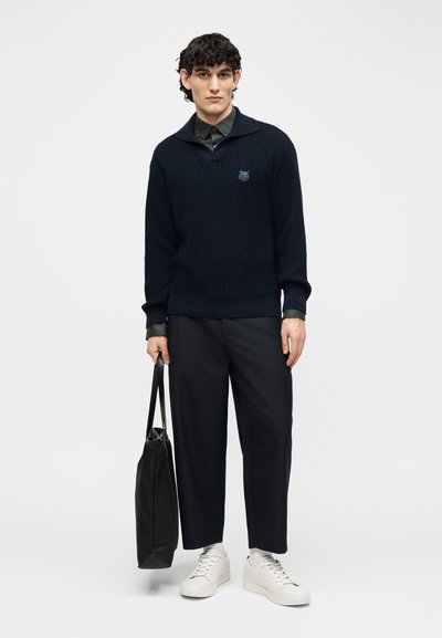 Homme portant un pull en tricot noir, un pantalon foncé, des baskets blanches, tenant un sac fourre-tout noir, debout devant un fond clair uni.