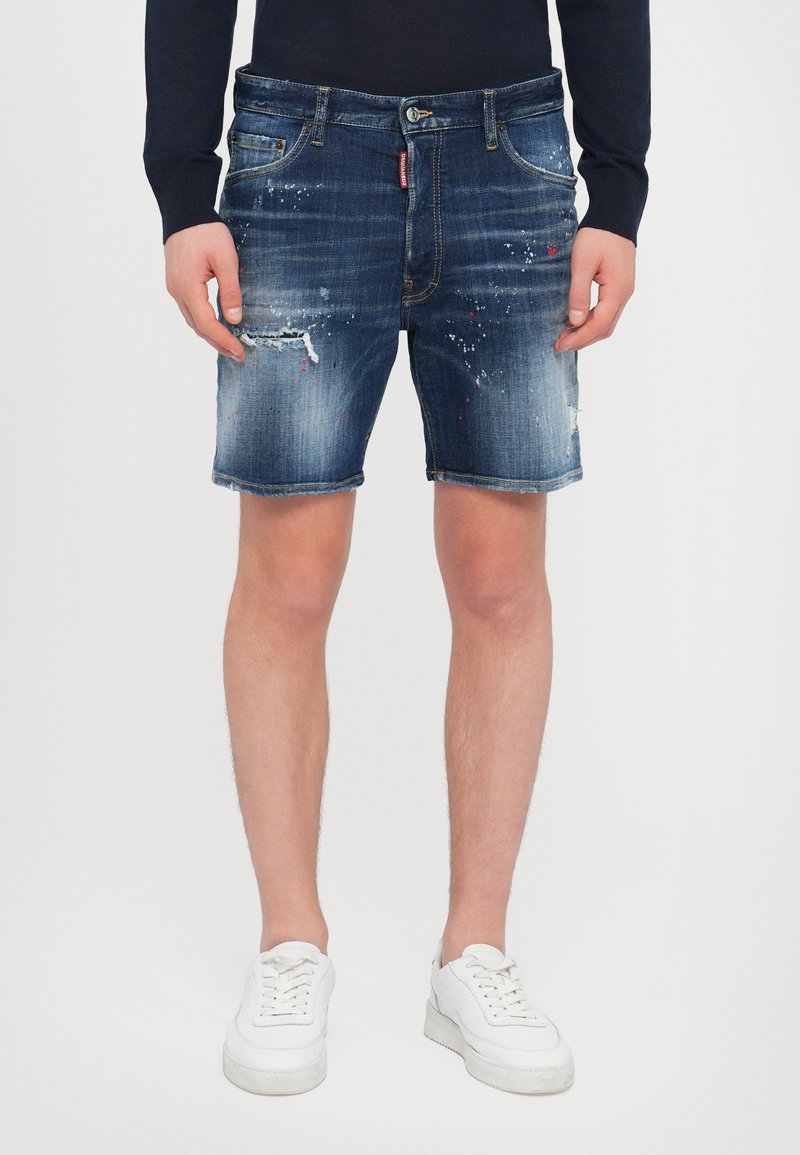 Dunkelblaue Denim-Shorts mit ausgefransten Kanten, abgenutzten Stellen und Farbspritzern. Kombiniert mit weißen Sneakern für einen lässigen Look.