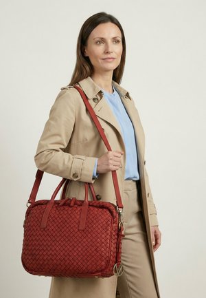 Žena v béžovém trenčkotě a světle modrém topu nese velkou červenou tkanou kabelku s ramenním popruhem na neutrálním pozadí.