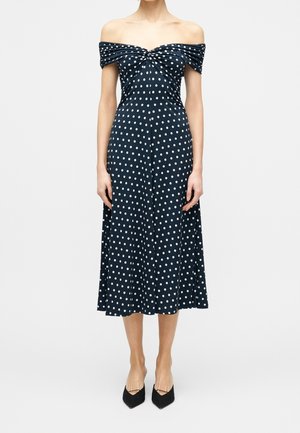 Kvinde iført en mørkeblå off-shoulder midi-kjole med hvide prikker og sorte pumps med spids tå, stående mod en ensfarvet baggrund.