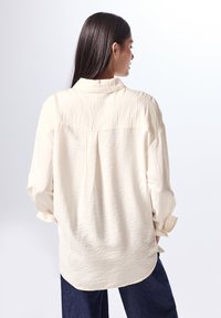 Camisa de textura crema con un corte relajado, mangas largas y cuello. Yugo trasero visible y dobladillo redondeado. Combinada con jeans azul oscuro.