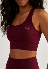Burgundowy stanik sportowy o teksturowanym wzorze, z małym białym logo serca po lewej stronie oraz dopasowanym, wspierającym kroju.