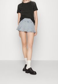 Schwarzes T-Shirt mit Nieten-Details, kombiniert mit einem hellblauen, plissierten Jeansrock. Weiße knielange Socken und schwarze Schuhe mit klobigem Absatz vervollständigen den Look.