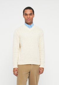 Homem com um suéter de malha creme sobre uma camisa azul de colarinho e calças de veludo cotelê bege, em pé contra um fundo branco.