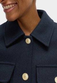 Marineblauer Wollmantel mit klassischem Kragen, goldenen Knöpfen mit dekorativem Emblem und zwei aufgesetzten Taschen auf der Vorderseite. Weiche Textur sichtbar.
