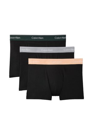 Atletyczny czarnoskóry mężczyzna stoi boso na szarym tle, nosząc czarne bokserki Calvin Klein, ręce za plecami.