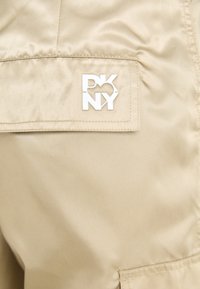 Beige nylonová tkanina s nášivkou bílého loga "DKNY." Textura je hladká, s pečlivě ušitými okraji a obdélníkovým tvarem.