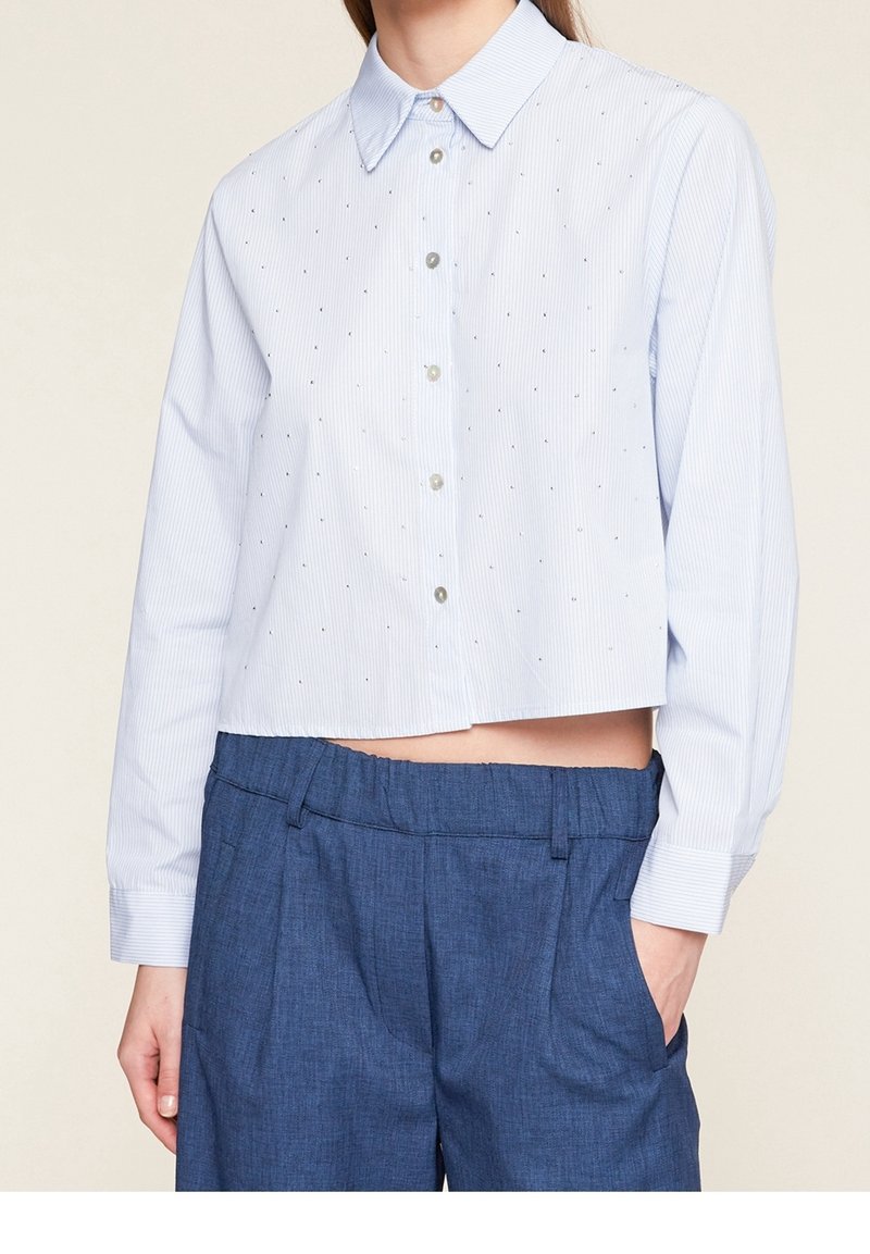 Femme vêtue d'une chemise courte bleu clair à manches longues avec de petits pois et d'un pantalon taille haute bleu foncé avec une ceinture élastique.