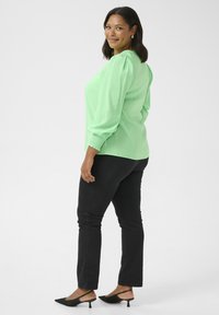 Blouse verte à manches longues avec des épaules bouffantes, associée à un jean slim noir et des escarpins à bout pointu noirs avec bride au talon.