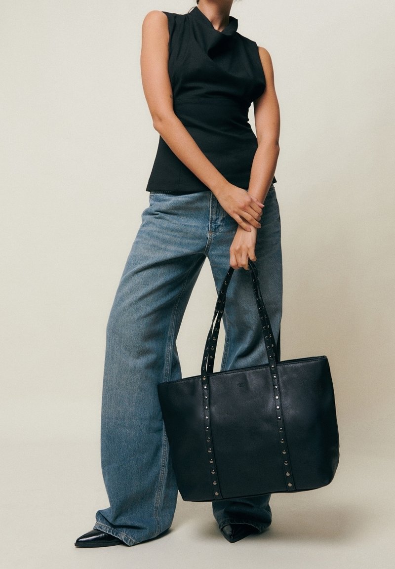 Femme portant un haut noir sans manches et un jean à jambes larges, tenant un grand sac cabas en cuir noir avec des sangles cloutées.
