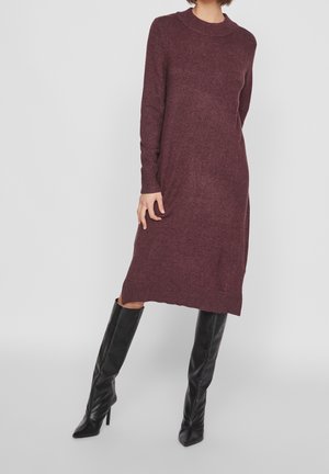 Vrouw in een pruimkleurige, knielange gebreide jurk met lange mouwen, gecombineerd met zwarte, kniehoge laarzen met een spitse neus en hak, tegen een effen achtergrond.